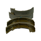 Close-up of two pairs of brake shoes — top pair showing curved grey friction lining with 12345 stamping, bottom pair showing gold zinc-plated steel webs with rivet holes and adjuster cut-outs