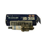 Part removed from box lying in front of open packaging; white sticker on box reads 'Bosch WR5AC Spark plug' with barcode; plug clearly shows white ceramic insulator with two blue stripe markings, hexagonal metal shell, and threaded section